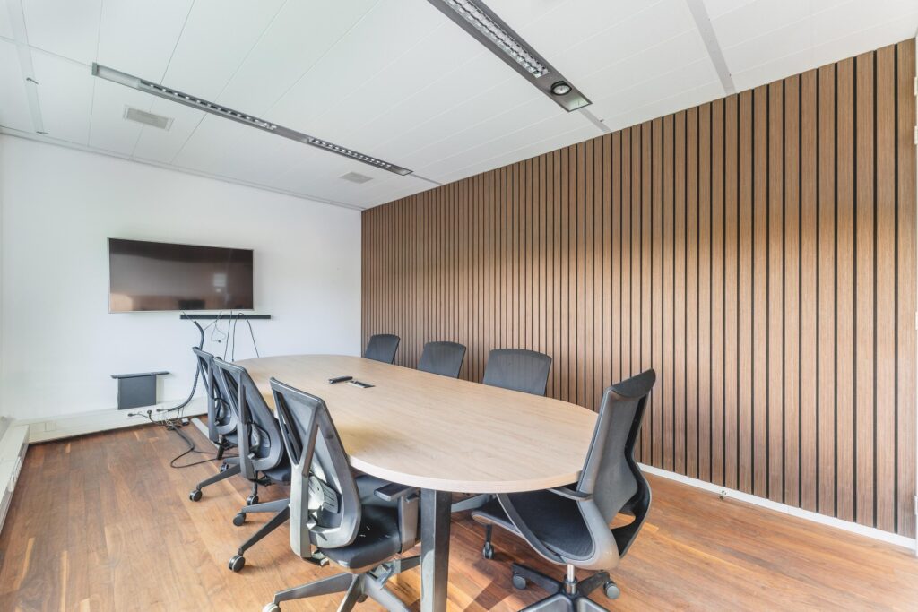 Vergaderruimte aan de Prof. W.H. Keesomlaan met ovale tafel, acht zwarte stoelen, houten lamellenwand en een flatscreen aan de muur.